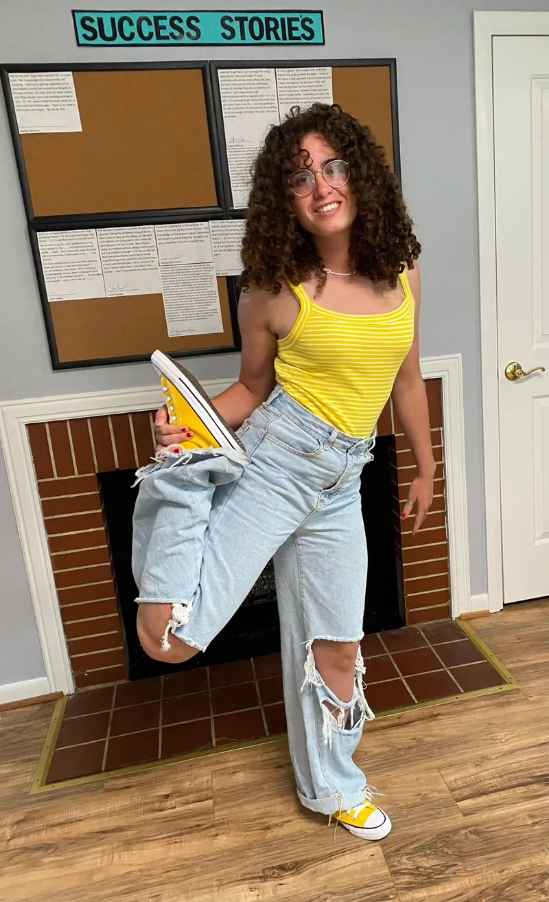 A relaxed, smiling young woman stretching one leg up with ease in front of the Success Stories board at Better Life Chiropractic in Groton, CT